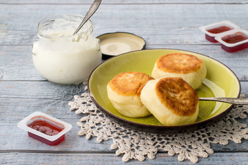 Delicious fried syrniki on a round plate, next to a glass jar with sour cream and small plastic containers with berry jam.