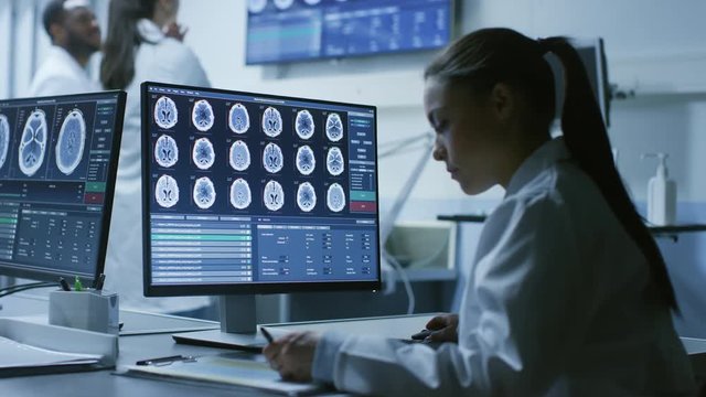 Female Medical Research Scientist Working with Brain Scans on Her Personal Computer, Writing Down Data in a Clipboard. Modern Laboratory Working on Neurophysiology, Science,  Neuropharmacology. 
