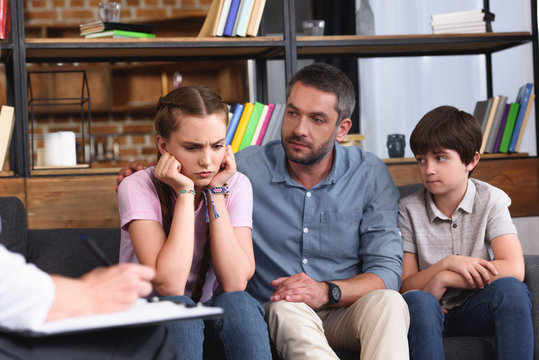 cropped image of female counselor writing in clipboard while man cheering up angry daughter on therapy session