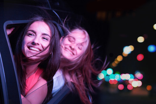 Two girls are riding in car and looking outside. THey are smiling and enjoying the moment. Their hair is waving by wind.