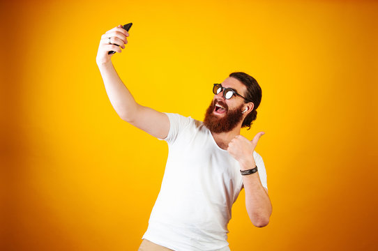 Young Cheerful man taking a selfie on his smartphone wearing a white T-shirt and earpods. Bearded long haired hipster looking at his iphone taking picture and showing thumb up on yellow background.
