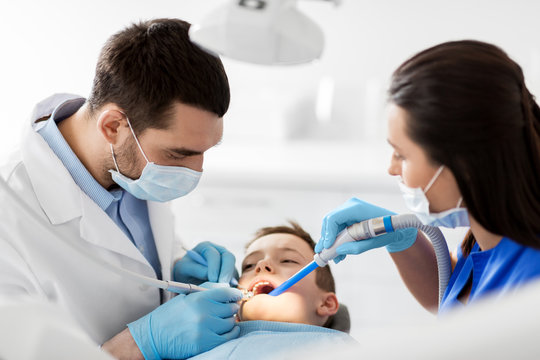 medicine, dentistry and healthcare concept - dentist and assistant with dental drill and saliva ejector treating kid patient teeth at dental clinic