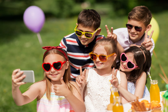 holidays, childhood and technology concept - happy kids in sunglasses taking selfie on birthday party at summer garden