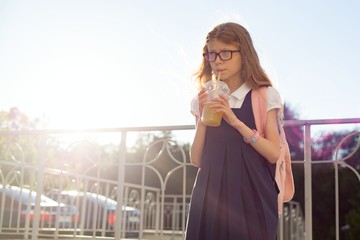 Outdoor portrait of girl elementary school student wearing glasses, school uniform, with backpack drinking natural juice from glass. Copy space.