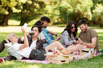 friendship, leisure, technology and people concept - group of friends with smartphones chilling on picnic blanket and drinking non alcoholic beer at summer park