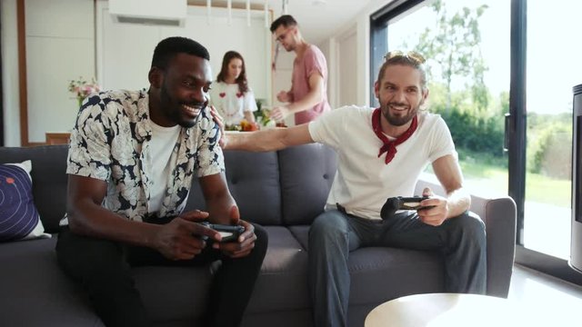 Two cheerful men joyfully play Xbox while sitting on a couch in a light living room, smiling couple preparing drinks on the background. True friendship, having fun, little party.