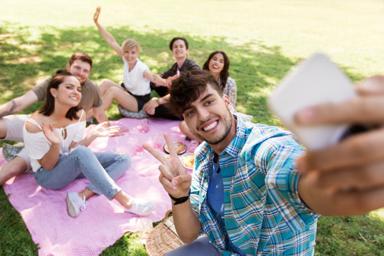 friendship, leisure and technology concept - group of happy smiling friends taking selfie by smartphone chilling on picnic blanket at summer park
