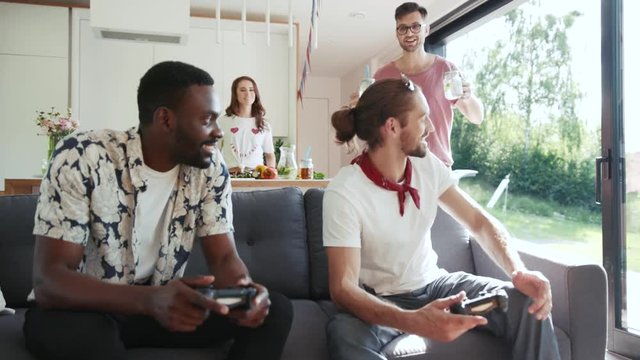 Multiracial company of friends playing Xbox, handsome young man in trendy glasses brings home made fresh lemonade in mason jars to them. Laughing, happy together, friendship.