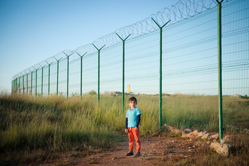 one little boy refugee in empty desert field near long fencing with barbed wire