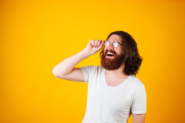 Happy young bearded long haired man trying glasses. Man wearing white t-shirt and round framed glasses on yellow background.