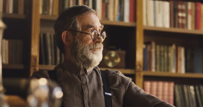 Portrait of the senior Caucasian professor in glasses sitting in his cabinet or library room with serious face.