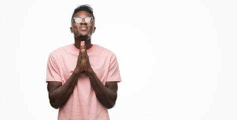 Young african american man wearing pink t-shirt praying with hands together asking for forgiveness smiling confident.
