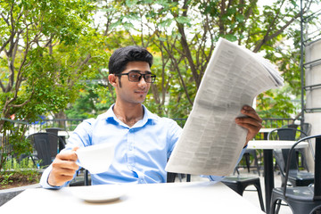 Positive business analyst drinking coffee and reading news. Young Indian in glasses looking through newspaper at outdoor cafe. Business and morning concept