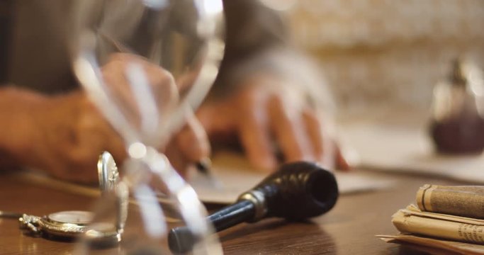 Close up of the different old things lying on the wooden table and blurred old male hands writing a letter with ink on the background.
