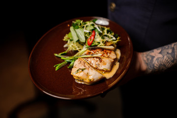 Tattoed bartender holding a plate of chicken fillet and salad