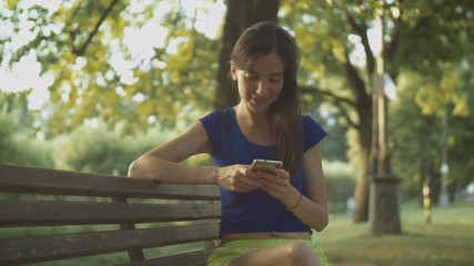 Cheerful beautiful long brown hair woman text messaging on smart phone while sitting on park bench in rays of setting sun. Lovely female networking with cellphone in park at sunset. Slow motion. - Powered by Adobe