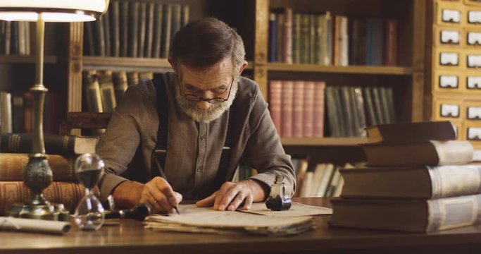 Portrait shot of the senior Caucasian man writing a letter and wetting a pen in the ink and starting to write on the old wooden table in the library room.