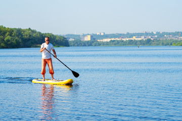 Naklejka premium Man sails on a SUP board in large river on the cityscape background