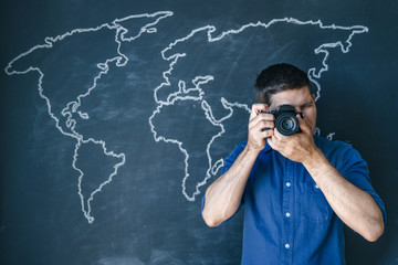 young man in a blue shirt on a background of a world map drawn in chalk on a black wall holds a camera in his hands, and takes a photo. The concept of travel and photography anywhere in the world.