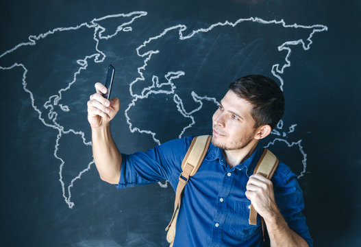 Man in a blue shirt with a yellow backpack and a smartphone on a wall background with a world map. Travel and mobile Internet.