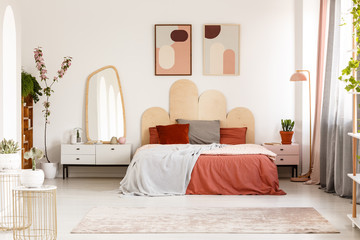 Modern posters above bed with headboard in pastel bedroom interior with mirror. Real photo