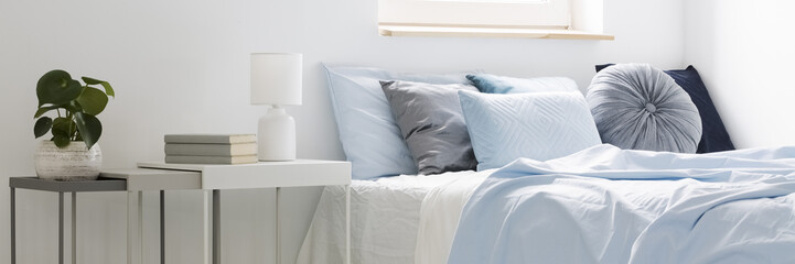 Real photo of a bed with blue bedding and cushions standing next to white tables with books, lamp...