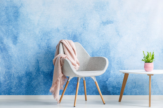Single armchair with pastel blanket standing next to a coffee table with a small plant on top against a blue and white ombre wall in a minimal sitting room interior. Copy space. Real photo.