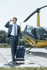 Satisfied with flight. Charming upbeat executive leaving the helipad having taken a helicopter flight while adjusting his eyeglasses and carrying a suitcase