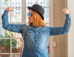 Stylish redhead woman wearing bowler hat and sunglasses showing arms muscles smiling proud. Fitness concept.
