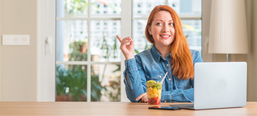 Redhead woman using computer laptop eating fruit at home very happy pointing with hand and finger...