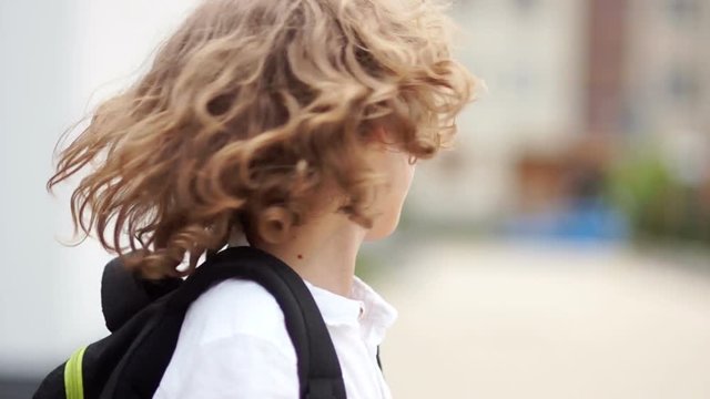 The blue-eyed boy corrects the harnesses of the backpack on his shoulder and throws back curly hair. Close up portrait, back to school, happy schoolboy