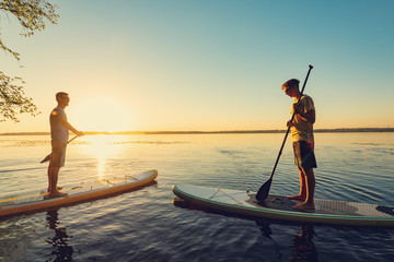 Guys, friends sail on a SUP boards in a rays of rising sun