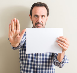 Senior man holding blank paper sheet with open hand doing stop sign with serious and confident expression, defense gesture