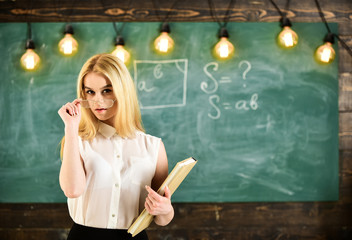 Teacher looks confident in eyeglasses, stand in classroom, chalkboard on background. Attractive lecturer concept. Woman with book starts lesson, gazes at audience while taking off eyeglasses.