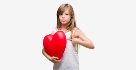 Young blonde toddler holding a red heart with surprise face pointing finger to himself