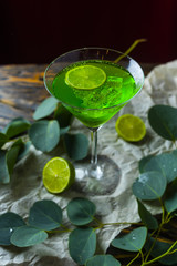 A green drink with ice cubes in a glass for mojito on crumpled wrapping paper next to which lie slices of lime and a branch of green tropical leaves on a vintage wooden table.