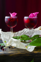 Two glases full of red drink with berries, ice and a pink flowers on crumpled wrapping paper beside which lie berries and a branch of green tropical leaves on a vintage wooden table.