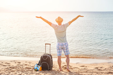 Man tourist in summer clothes with a suitcase in his hand, looking at the sea on the beach, concept of time to travel