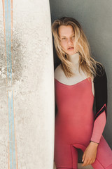 portrait of attractive young woman in wetsuit with surfing board standing against grey wall
