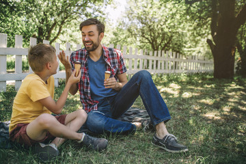 Naklejka na ściany i meble Happy father is sitting besides his son on blanket and smiling to him. Both he and child have ice creams. Guys are enjoying spending time together.