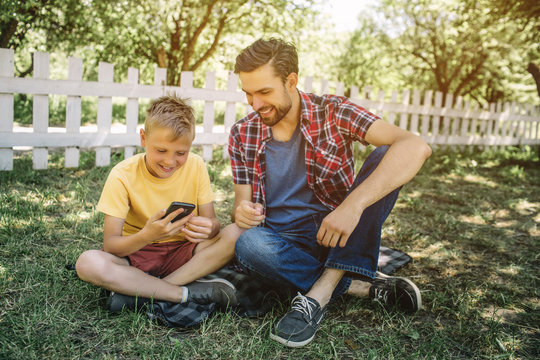 Small boy is sitting on grass with his dad with their legs crossed. Child is holding phone in hands and looking at it. He is smiling. Dad is looking at device too and smiling as well.