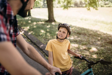 Obraz premium Nice picture of cheerful child looking up on his father. He is standing at his bicycle. Adult guy is standing besides his son and looking at him.