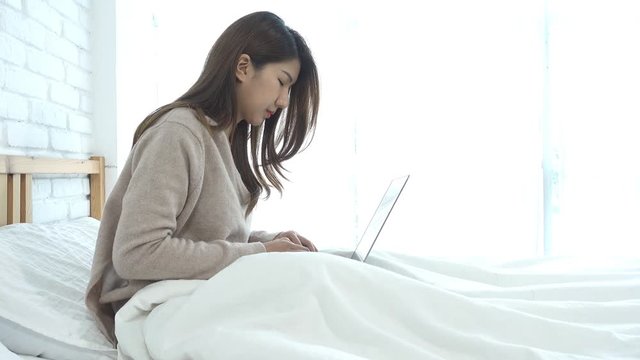 Beautiful young smiling asian woman working on laptop while sitting on bed in bedroom at home. Asian business woman relax in her home. Enjoying time at home.