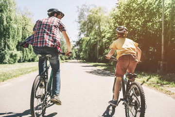 A picture from the back. Man and child are riding on bicycles together on one road. They have a compatition. Right now father is winning.