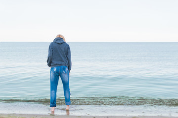 A woman dressed in jeans and a sweater stands on the seashore in the water. Concept Women's loneliness, depression.