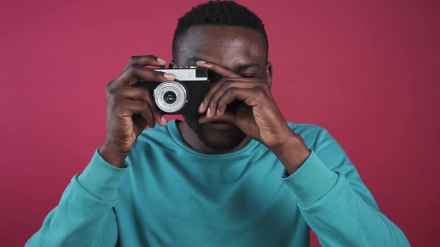 Portrait of good-looking African guy in colored sweater. Nice young photographer smiling, taking photos on background of pink wall. Close-up. Shiny white smile.