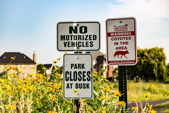 Sign warning of coyotes in the area, no motorized vehicles & park closes at dusk in a suburban area. Houses are visible in the distance. Concepts of wildlife, human wildlife conflicts and interactions