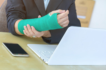 injured woman with broken hand sitting and holding green cast working with laptop in office