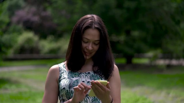 Happy looking asian woman walks in the park and types sms on the yellow smartphone.