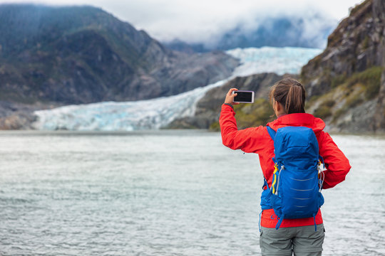 Alaska travel cruise ship tourist taking photo with phone. Hiker with backpack at Mendenhall glacier in Juneau visiting famous attraction destination. Glacier arm melting in lake.